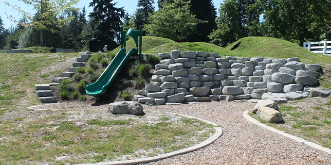 short slide beside a rock wall at big rock park north, with small grass hillocks at the top of the slide and stairs to access